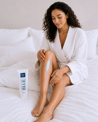 A woman in a white bathrobe sits on a white bed applying light blue sunscreen cream to her leg, with a tube of Blue Spectrum sunscreen standing nearby.
