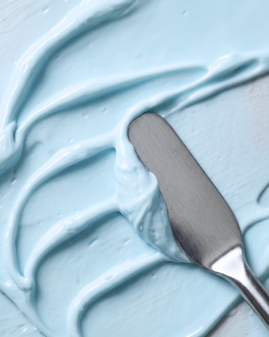 A close-up of light blue whipped cream or frosting swirled on a surface with a metal spatula resting on top.